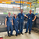 Zwei Frauen und zwei Männer stehen vor einer Anlage, gekleidet in blaue Arbeitskleidung.