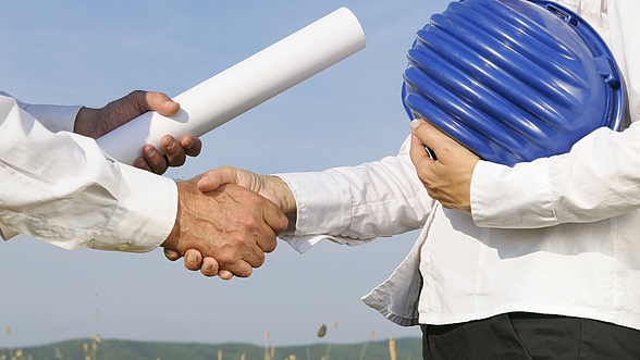 Zwei Männer schütteln sich die Hände. Der eine hat einen Arbeitshelm unter dem Arm und erhält ein Zertifikat.