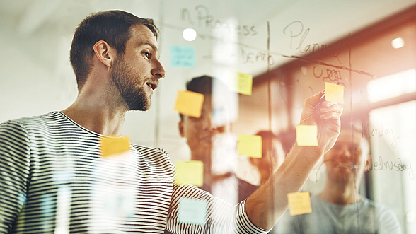 © PeopleImages -istock.com  Ein Mann steht an einer Wand und beschriftet Klebezettel.