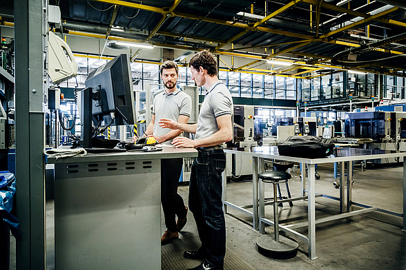 Zwei Männer stehen in einer Industriehalle vor einem Rechner und führen ein Gespräch.