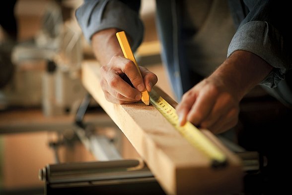 Hände, die mit einem Bleistift und Zollstock eine Markierung auf einem Stück Holz anbringen.