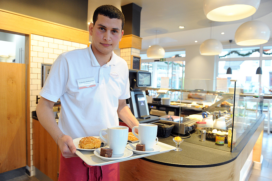 Zakaria Dahmoun mit einem Tablett mit Kaffee und Kuchen in der Hand.