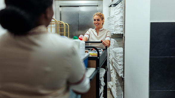 © Dimensions -istock.com  Zwei Reinigungskräfte schieben einen Wäschewagen durch einen Flur