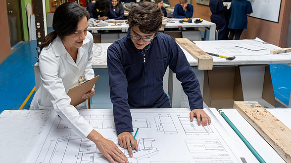 ©andresr - istock.com Eine Ausbilderin mit weißem Kittel zeigt einem Azubi auf einem Plan etwas.