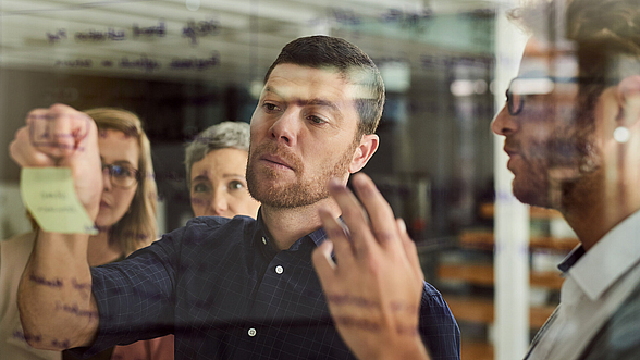 © Jeffbergen – istock.com  Mann plant an Scheibe mit Kolleginnen