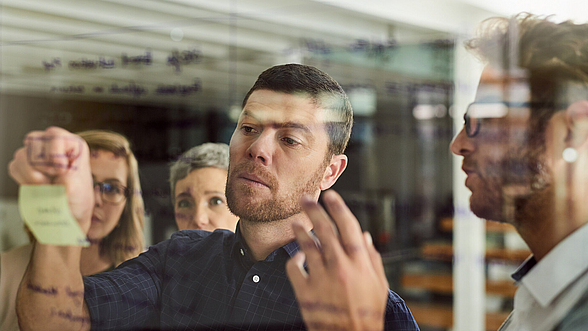 Ein Ausbilder klebt einen Klebezettel auf eine Glas-Pinnwand 