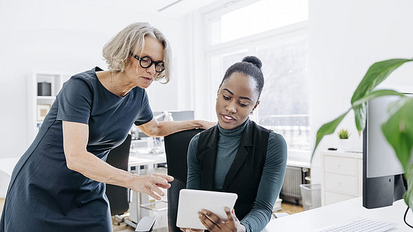 Seniorin im Gespräch mit jungem Kollegen mit digitalem Tablet bei der Arbeit