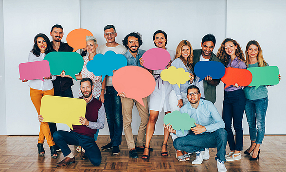 Zu sehen ist eine Gruppe von 12 Personen, die bunte Sprechblasen in der Hand halten. Es ist kein Text auf den Sprechblasen zu lesen.