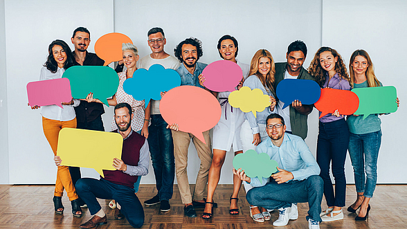 Zu sehen ist eine Gruppe von 12 Personen, die bunte Sprechblasen in der Hand halten. Es ist kein Text auf den Sprechblasen zu lesen.
