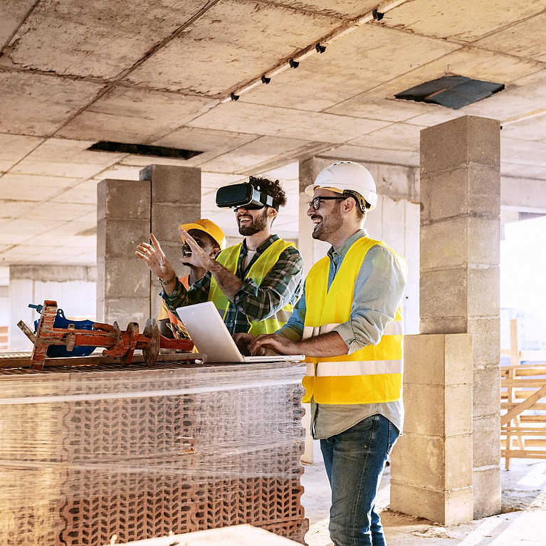 Drei Bauarbeiter nutzen Laptop und Virtual-Reality-Brille auf einer Baustelle