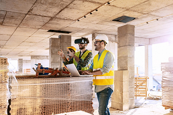 Drei Männer in gelben Warnwesten sehen sich in einem Rohbau die Zukunft mit VR-Brillen an