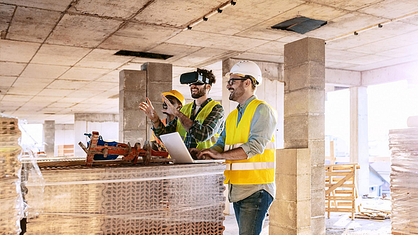 © ljubaphoto-istock Drei Männer in gelben Warnwesten sehen sich in einem Rohbau die Zukunft mit VR-Brillen an