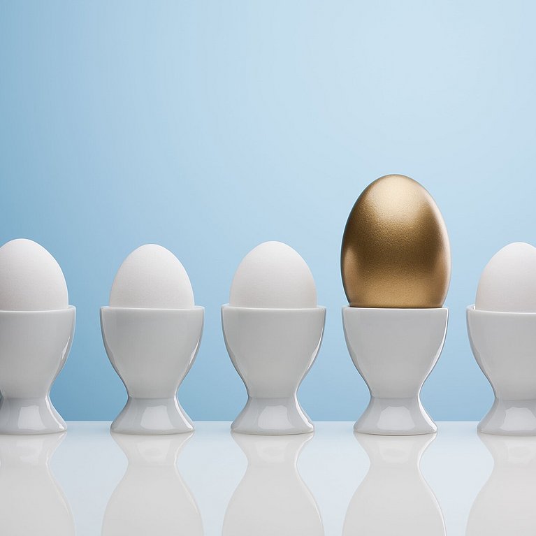 Zu sehen sind verschiedene Eier, die im Eierbecher aufgereiht sind. Ein Ei ist gold, alle anderen sind weiß.