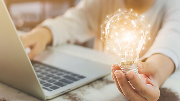 Person am Laptop, diese hält eine leuchtende Glühbirne in der Hand
