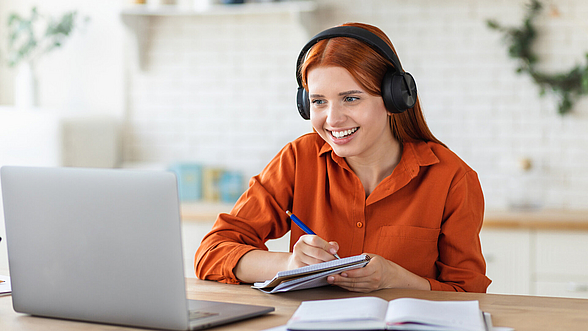 © KucherAV - istock.com Eine junge Frau sitzt lächelnd mit einem Headset und einem Notizblock vor einem Laptop.
