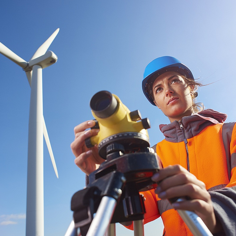 Auszubildende zur Vermessungstechnik steht mit Vermessungsgerät vor Windkraftrad