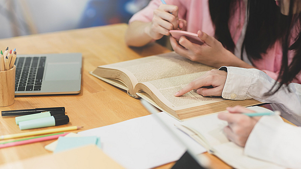 Laptop, Bücher und Papiere liegen auf einem Tisch, an dem zwei Personen arbeiten