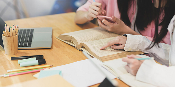 Laptop, Bücher und Papiere liegen auf einem Tisch, an dem zwei Personen arbeiten