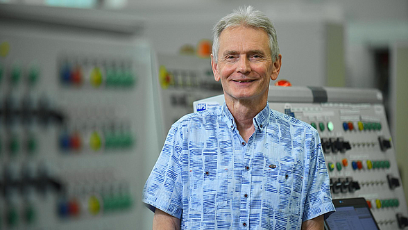 © aktiv/Christian Augustin  Älterer Mitarbeitender steht in einer Produktionsstätte und lächelt in die Kamera.