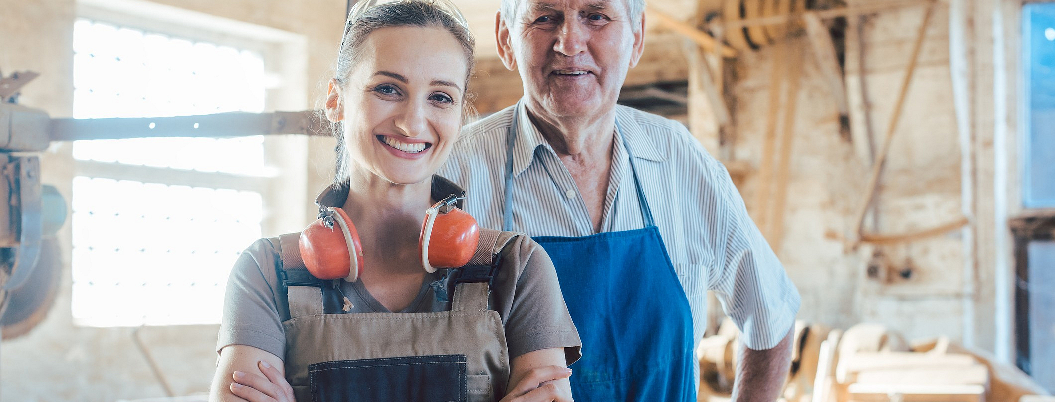 Senior-Schreinermeister mit seiner Enkelin in der Holzwerkstatt beim Blick in die Kamera.