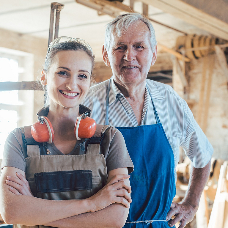 Senior-Schreinermeister mit seiner Enkelin in der Holzwerkstatt beim Blick in die Kamera.