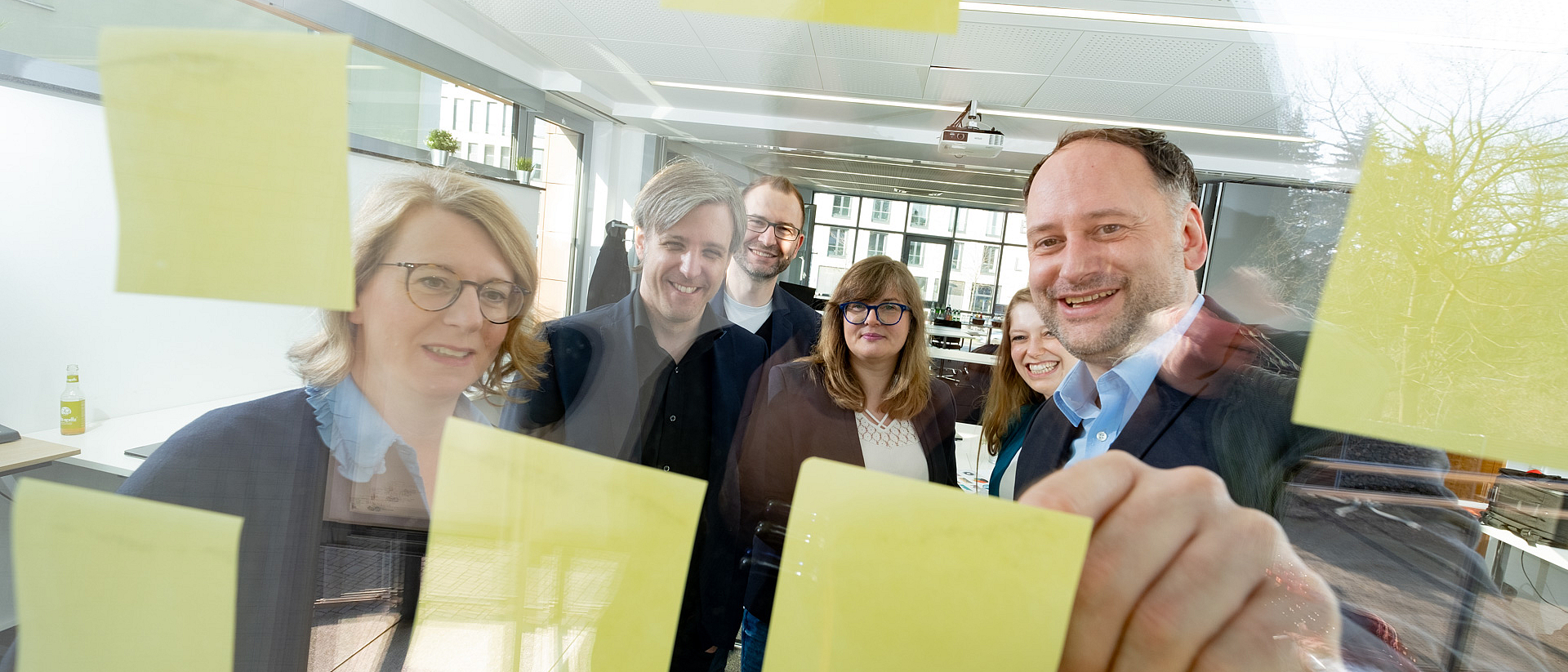 © dietrichkuehne_Arineo Mehrere Teammitglieder in einem Büro blicken positiv gestimmt auf ein Fenster, auf dem Post-Its kleben.