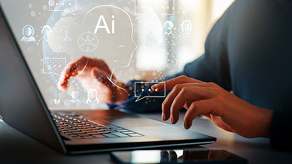 © Choreograph – istock.com Person tippt am Computer über dessen Tastatur eine wolkige Darstellung Technologie abbildet.