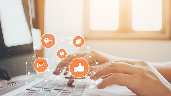 Zu sehen ist eine Hand, die auf einer Computer-Tastatur tippt. Im Vordergrund sind Symbole, die auf Social Media genutzt werden.