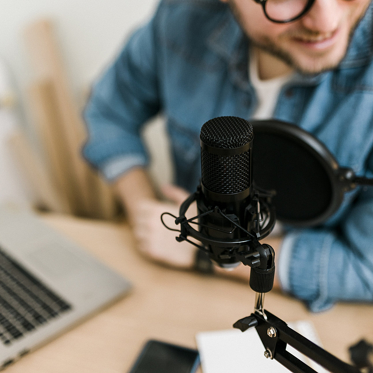 Ein mittelalter Mann sitzt vor einem Laptop und einem professionellen Aufnahmegerät für Podcasts.