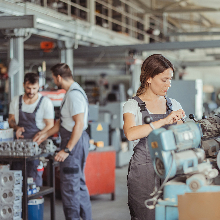 Arbeitssituation in einer Werkstatt. Junge Frau arbeitet an Maschine im Hintergrund arbeiten zwei Kollegen.