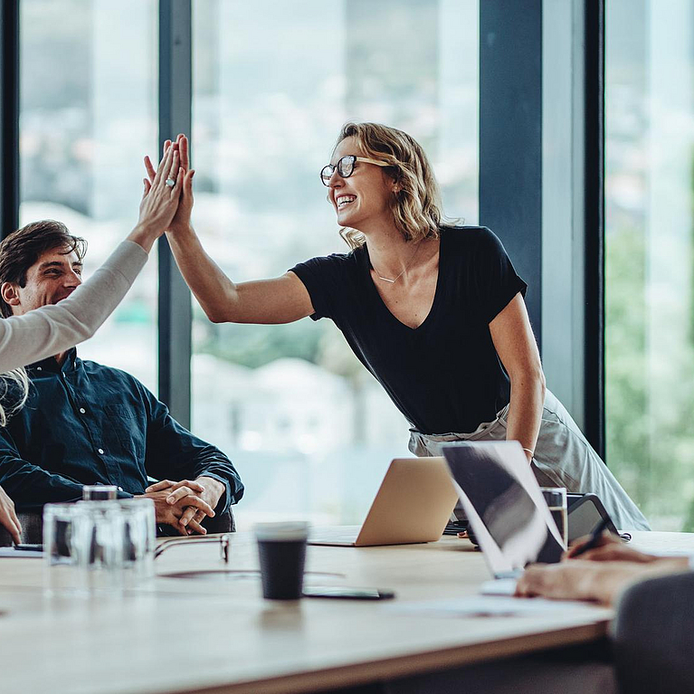 Mehrere Personen sitzen an einem Tisch. Zwei Personen geben sich High Five.