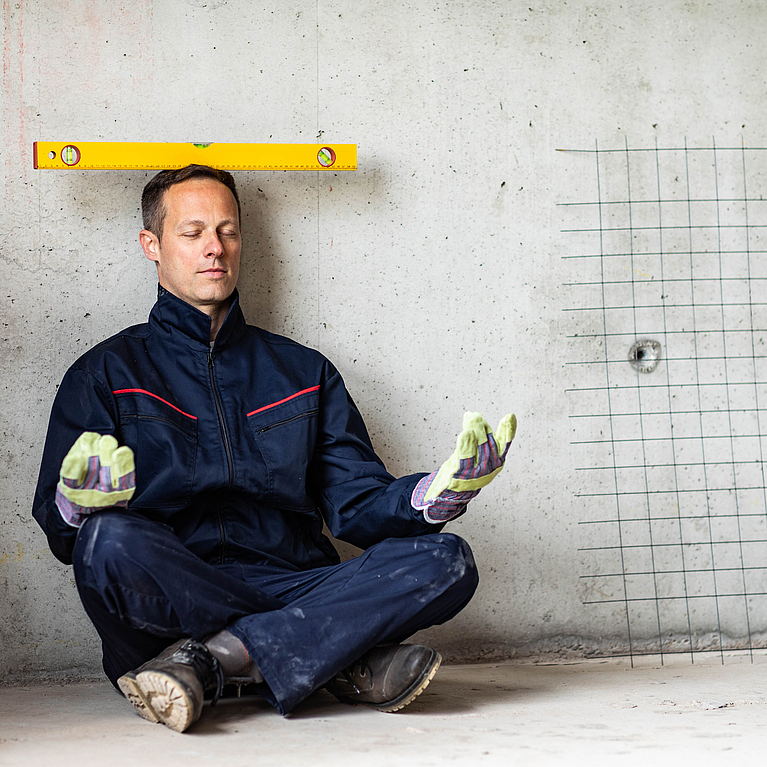 Ein Handwerker sitzt im Schneidersitz auf dem Boden und balanciert eine Wasserwage auf dem Kopf.